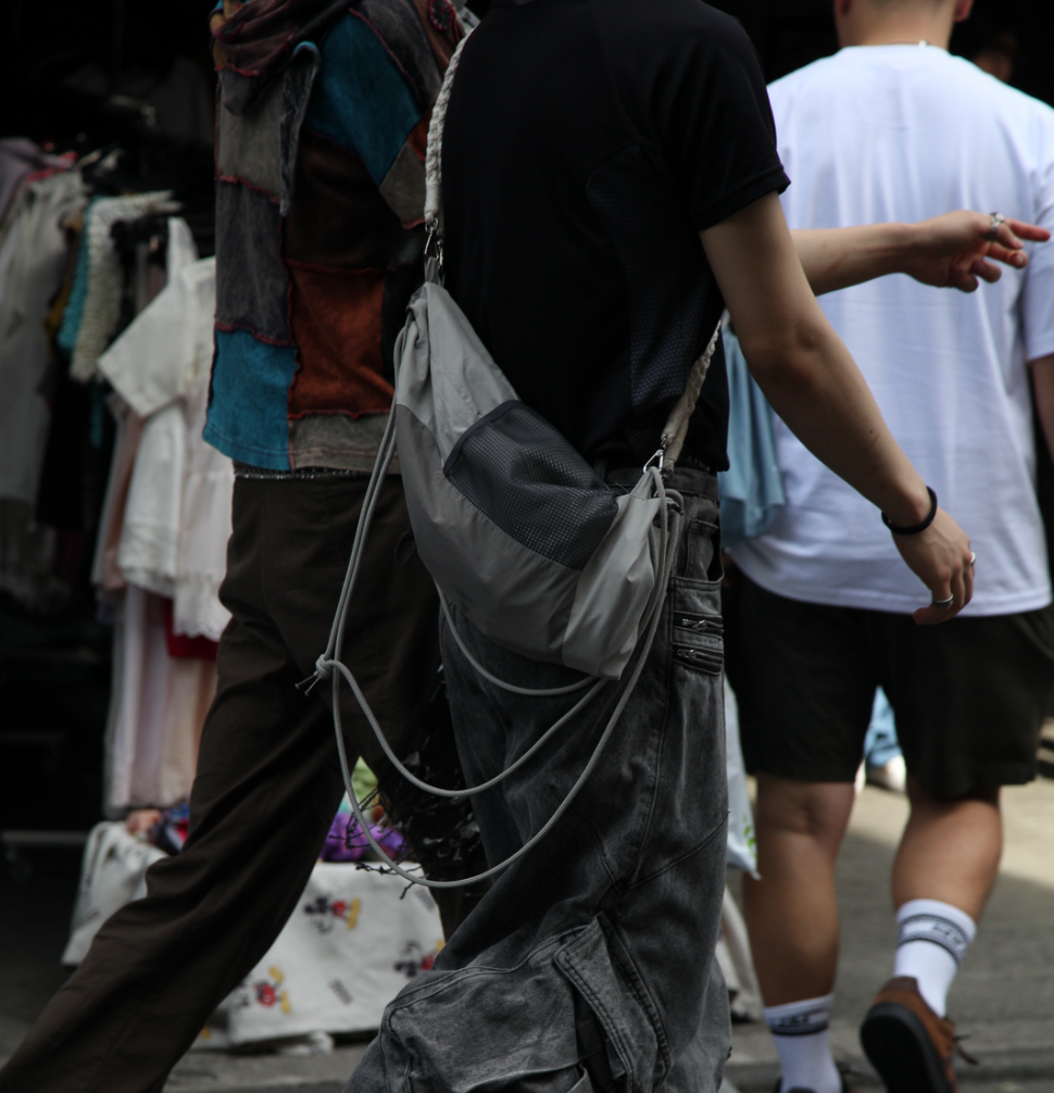 A street fashion scene in Japan featuring two men walking. The man in the foreground wears a gray crossbody bag with a braided strap and mesh pocket, coordinating with his dark shirt and loose-fitting cargo pants. His casual, yet carefully styled outfit emphasizes soft materials and functional design. The second figure wears a colorful patchwork jacket, adding a contrast of textures and colors to the scene. The background shows a clothing stall with hanging garments."