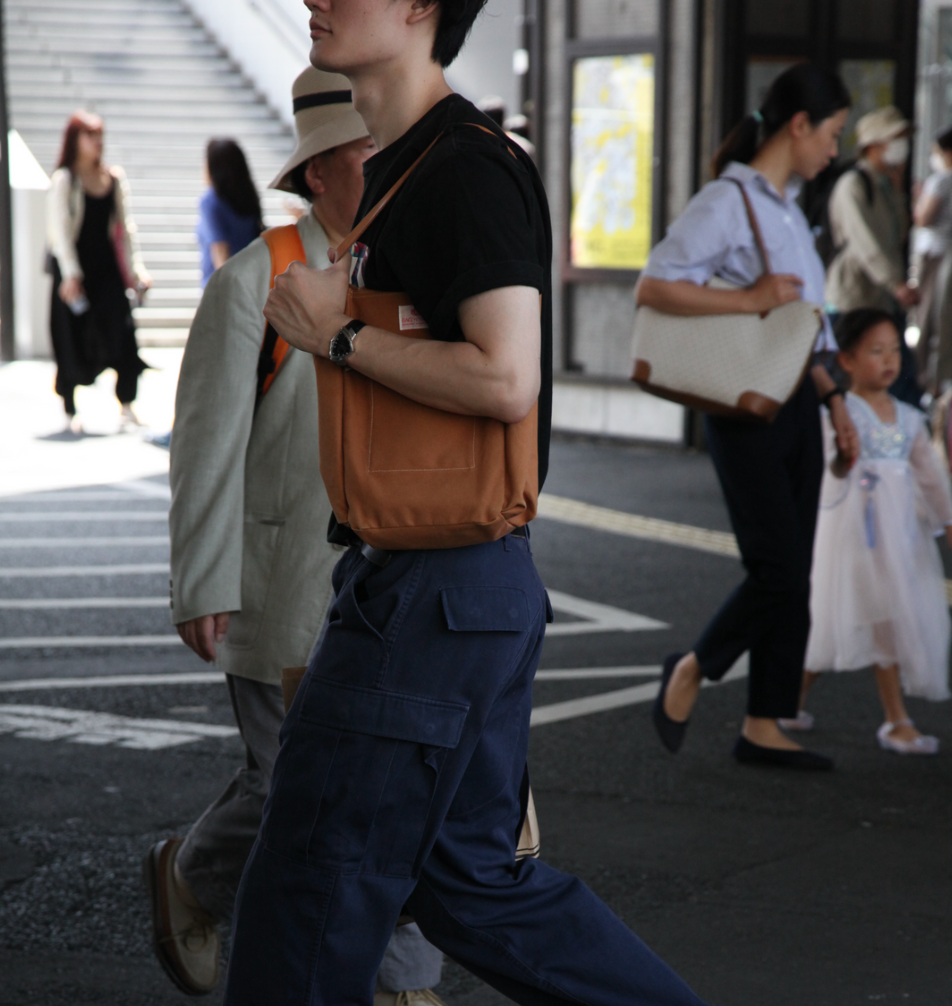 A man walking through a busy street in Japan, dressed in a casual outfit with a black t-shirt, navy cargo pants, and carrying a tan shoulder bag. His outfit is simple yet practical, with the bag slung casually over his shoulder. In the background, several people walk by, including a child in a blue dress holding hands with a woman. The scene captures a candid moment of everyday street style, blending casual and functional fashion.