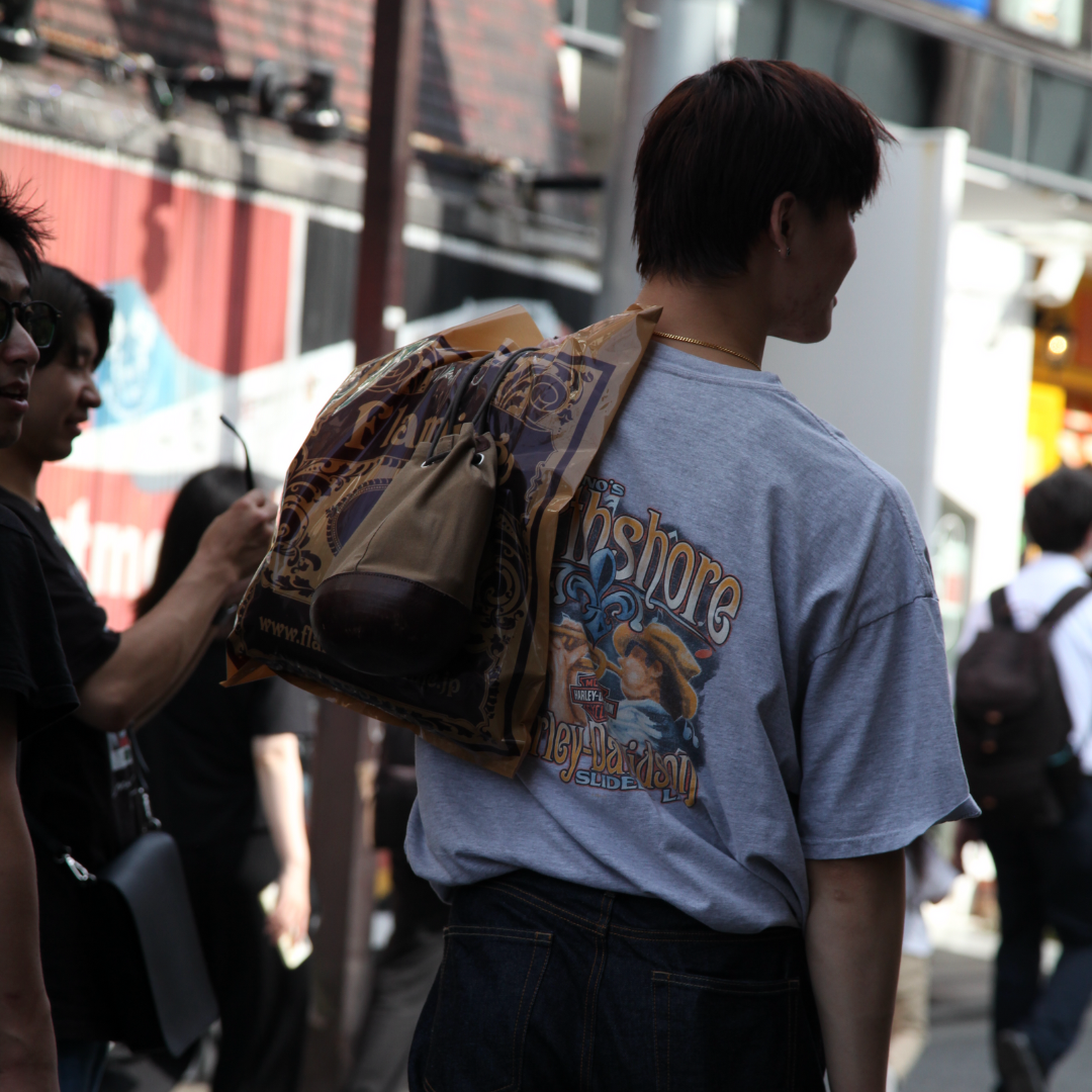 A young man wearing a gray t-shirt with a colorful design on the back, carrying a patterned bag over his shoulder, stands in a crowded urban setting with several people visible in the background.